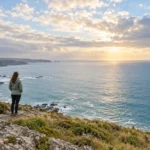 A woman stands on a quiet coastal overlook facing a wide sea and open sky.