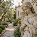 An aged stone angel sculpture stands among olive trees beside a quiet chapel garden path.